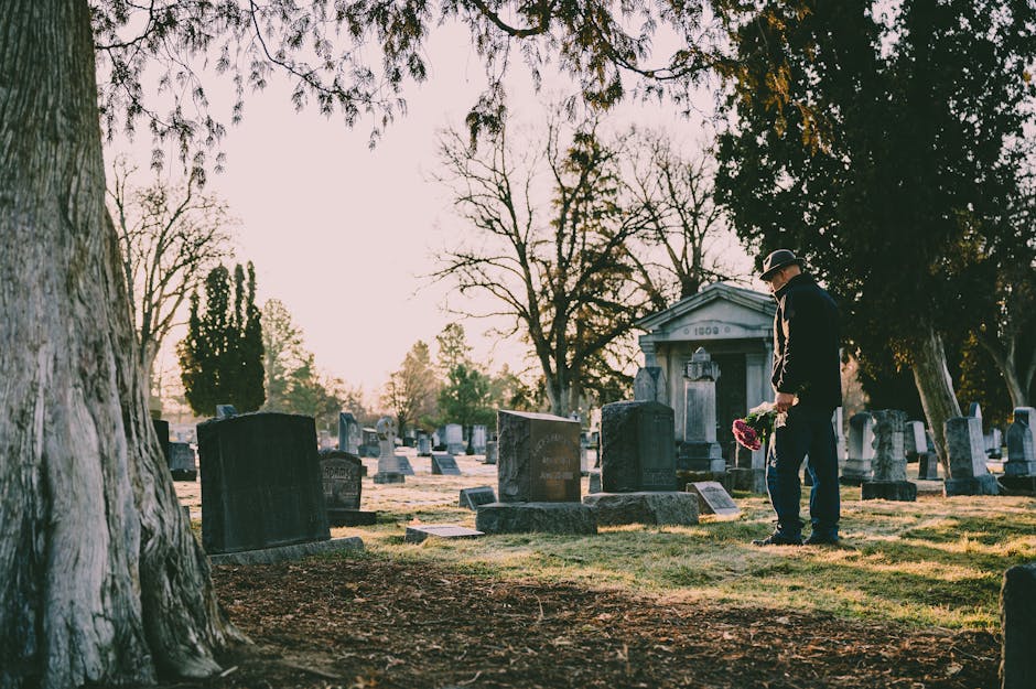 A man dressed in dark clothing and a hat stands in a cemetery holding a bouquet of pink and white flowers. The arrangement features a variety of fresh, vibrant blooms wrapped in clear cellophane, plac