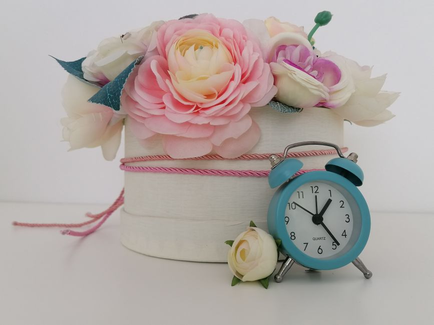 A floral arrangement of artificial flowers in pastel pink, white, and cream tones, including roses and peonies, set inside a white cylindrical hatbox-style container decorated with a pink braided ribb