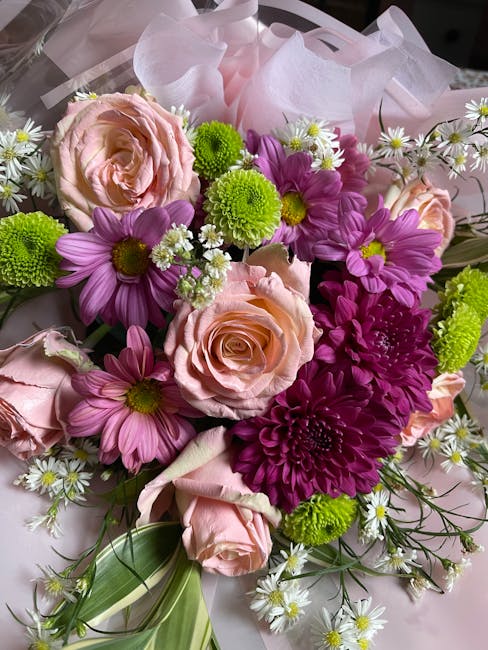 A vibrant floral bouquet featuring soft pink roses, purple daisies, and bright green chrysanthemums arranged together. The roses exhibit delicate, layered petals with a gentle sheen, while the daisies