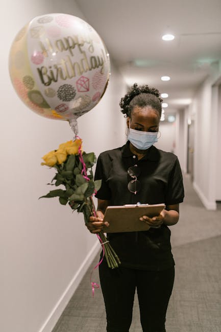 A woman wearing a light blue face mask and a black blouse is standing in a well-lit corridor, holding a bouquet of fresh yellow roses wrapped in green foliage and tied with a pink ribbon. She is also